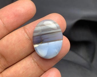May include: An oval-shaped gemstone with a polished surface. The stone features bands of gray, purple, and light blue hues. The gemstone is held in a hand, with a dark background.