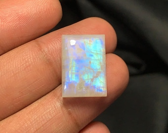 May include: A rectangular moonstone gemstone with a pearly white base and iridescent blue and green flashes. The stone is held in a hand, showcasing its polished surface and natural patterns. The gemstone is a cabochon cut.