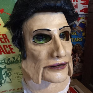 May include: A realistic-looking, hand-painted, white and flesh-toned, male mask with black hair. The mask has green eyes, a painted-on smile, and a painted-on purple lip. The mask is sitting on a white surface.