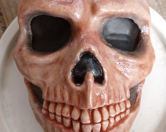 May include: A detailed, realistic skull mask with a fleshy, pale pink and red color scheme. The mask has a wide, toothy grin and black eye sockets.