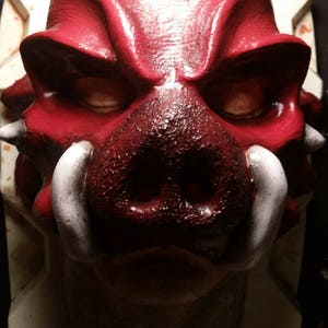 May include: A red and white boar mask with a textured surface and large tusks. The mask is mounted on a white background.