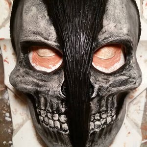 May include: A black and white skull mask with a black mohawk. The mask has a textured surface and is made of a hard material.