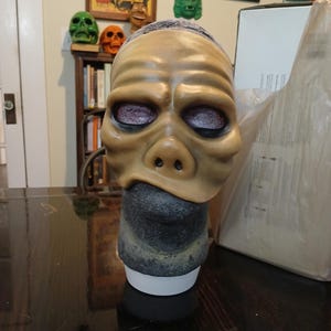 May include: A tan-colored latex mask with a large, flat nose and large, dark purple eyes. The mask is on a mannequin head with a dark gray, textured surface.