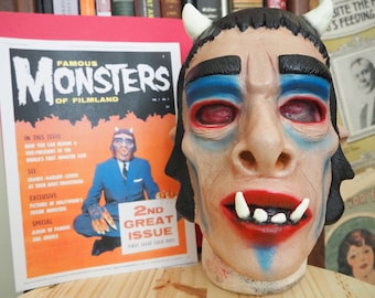 Può includere: Una maschera da mostro blu e rossa con corna, ombretto blu e una bocca rossa con denti bianchi. La maschera è su una superficie di legno davanti a una rivista intitolata "Famous Monsters of Filmland".