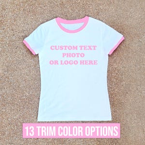 May include: A white ringer t-shirt with pink trim on the collar and sleeve cuffs. The center of the shirt features the pink text CUSTOM TEXT PHOTO OR LOGO HERE. Below the shirt, a pink rectangular banner displays the text 13 TRIM COLOR OPTIONS.