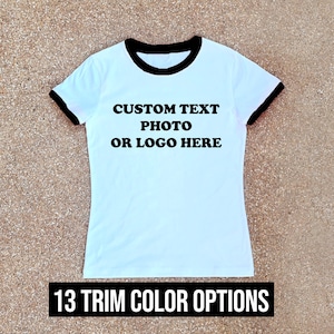 Op de afbeelding: Wit T-shirt met zwarte rand rond de kraag en mouwen. Het shirt heeft de tekst "CUSTOM TEXT PHOTO OR LOGO HERE" op de voorkant. De onderkant van de afbeelding heeft de tekst "13 TRIM COLOR OPTIONS".