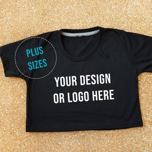 May include: Black cropped t-shirt with short sleeves and a round neckline. The shirt has the text "YOUR DESIGN OR LOGO HERE" printed on the front. The text "PLUS SIZES" is circled in blue on the left side of the shirt.