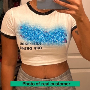 May include: White cropped t-shirt with black trim on the sleeves and neckline. The shirt features a blue and white graphic of a hand with the text "OFF DRUGS KEEP KIDS" printed on the front.