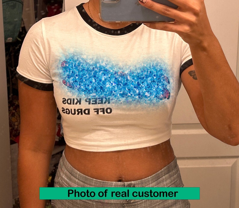 May include: White cropped t-shirt with black trim on the sleeves and neckline. The shirt features a blue and white graphic of a hand with the text "OFF DRUGS KEEP KIDS" printed on the front.