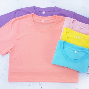 May include: A stack of four plain t-shirts in different pastel colors: light purple, peach, yellow, and light blue. The shirts are folded and arranged on a white marble surface.