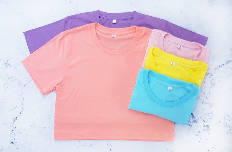May include: A stack of four plain t-shirts in different pastel colors: light purple, peach, yellow, and light blue. The shirts are folded and arranged on a white marble surface.