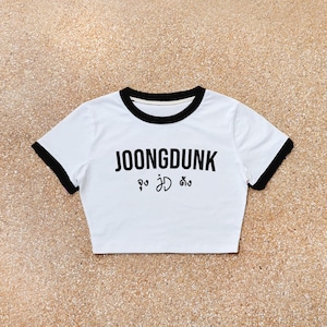 Puede incluir: Top corto blanco con ribete negro en el cuello y las mangas. El texto "JOONGDUNK" está impreso en negro, junto con texto adicional en una fuente diferente. Estilo casual.