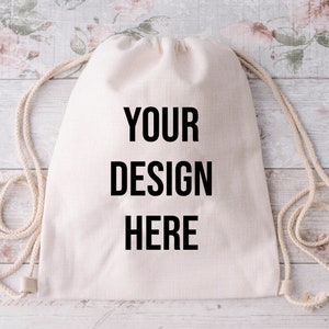 Op de afbeelding: Een witte trekkoordtas met de tekst "YOUR DESIGN HERE" in het zwart gedrukt op de voorkant.