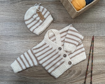 May include: A cream-colored, striped baby sweater and matching hat set. The sweater has three brown buttons and a V-neck. The hat has a small tag that says "handmade". The set is made of a soft, knitted material. Knitting needles and yarn are in the background.