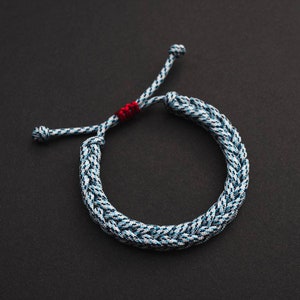 May include: A braided rope bracelet with a blue and white pattern and a red accent. The bracelet is on a black background.