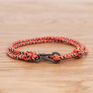 May include: A bright orange and black braided rope bracelet with a black metal clasp. The clasp has the letters "SF" engraved on it.