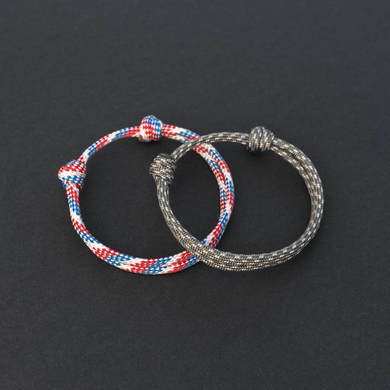 May include: Two braided rope bracelets, one with red, white, and blue stripes and the other with a gray and black pattern.