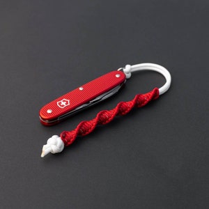Puede incluir: Una navaja de bolsillo roja con un logotipo de cruz blanca unida a un cordón trenzado rojo y blanco.