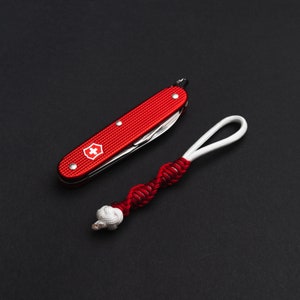 Op de afbeelding: Een rood zakmes met een wit kruislogo en een rood-wit gevlochten koord.