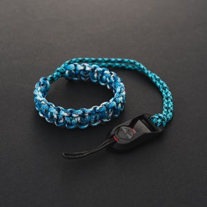 Op de afbeelding: Een blauwe en witte paracord camera polsband met een zwarte verstelbare gesp en een rode accent.