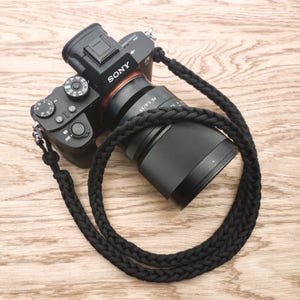 Könnte beinhalten: Eine schwarze Kamera mit einem schwarzen geflochtenen Kameragurt. Das Kameraobjektiv ist ein Sony FE 50mm f/1.8 Objektiv.