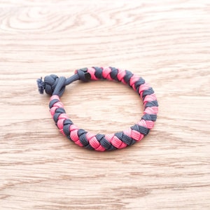 May include: A pink and gray braided paracord bracelet. The bracelet is made with a knot at the end.