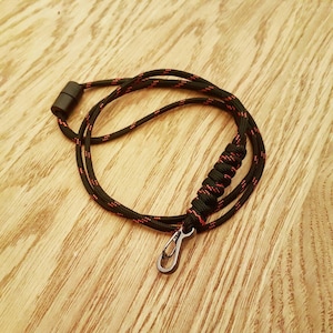 Könnte beinhalten: Ein schwarzes und rotes Paracord-Lanyard mit einem Metallclip. Das Lanyard besteht aus einem geflochtenen Seil mit roten Akzenten.