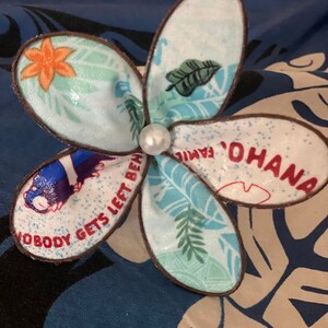 Puede incluir: Un pin de tela en forma de flor azul y blanco con ribete marr&oacute;n. El pin tiene una perla en el centro y presenta el texto "Ohana" y "Nobody gets left behind."