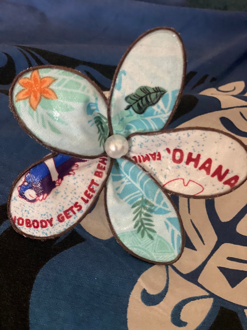 Puede incluir: Un pin de tela en forma de flor azul y blanco con ribete marr&oacute;n. El pin tiene una perla en el centro y presenta el texto "Ohana" y "Nobody gets left behind."