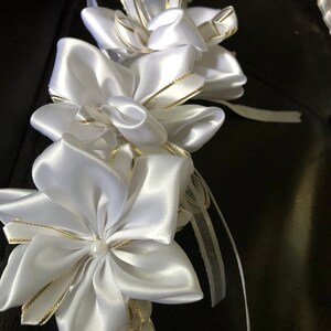 May include: A white satin lei with gold trim and a pearl center. The lei features multiple flower-shaped accents and a braided ribbon design. The background is a dark surface.