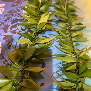 May include: A green lei made of woven leaves, with a dark blue and white patterned fabric in the background.