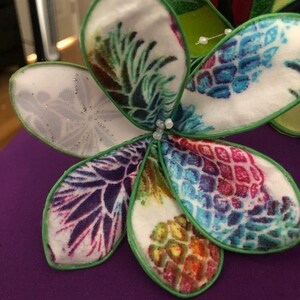 Puede incluir: Una flor de tela blanca con bordes verdes y un estampado de pi&ntilde;a. La flor tiene cinco p&eacute;talos y est&aacute; decorada con peque&ntilde;as cuentas blancas.