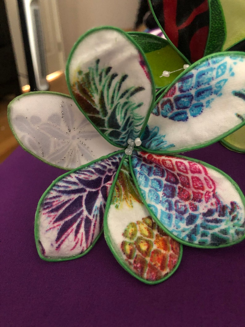Puede incluir: Una flor de tela blanca con bordes verdes y un estampado de pi&ntilde;a. La flor tiene cinco p&eacute;talos y est&aacute; decorada con peque&ntilde;as cuentas blancas.