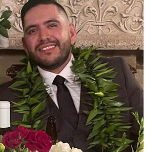 May include: A man in a black suit and a green lei smiles at the camera. He is wearing a black tie and has a bottle of wine and a bottle of beer in front of him. There are red and white flowers in the foreground.