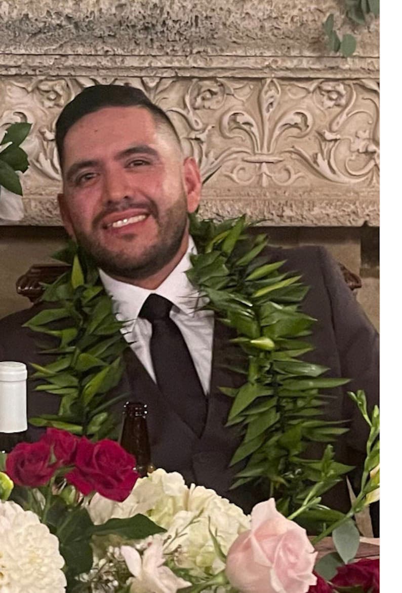 May include: A man in a black suit and a green lei smiles at the camera. He is wearing a black tie and has a bottle of wine and a bottle of beer in front of him. There are red and white flowers in the foreground.