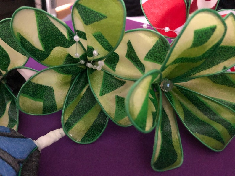 Puede incluir: Primer plano de una flor verde y amarilla hecha de tela con cuentas blancas y una pedrer&iacute;a transparente. La flor est&aacute; sobre una superficie morada.