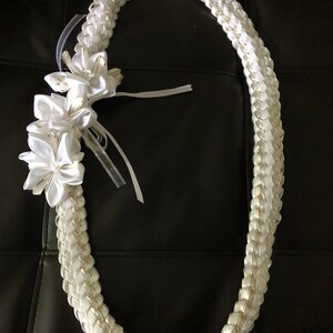 May include: A white satin lei with a braided design, accented with gold trim. The lei features three white flower accents and sheer white ribbons. The lei is a traditional Hawaiian garland, often worn for celebrations and special occasions.