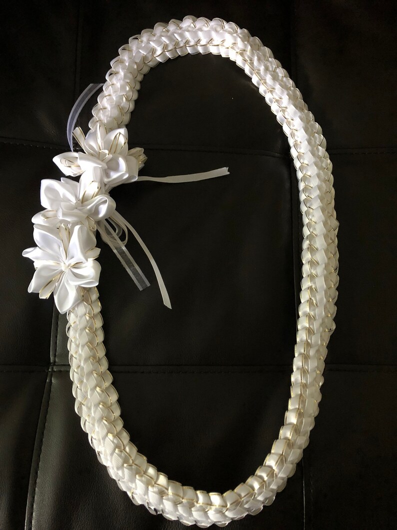 May include: A white satin lei with a braided design, accented with gold trim. The lei features three white flower accents and sheer white ribbons. The lei is a traditional Hawaiian garland, often worn for celebrations and special occasions.