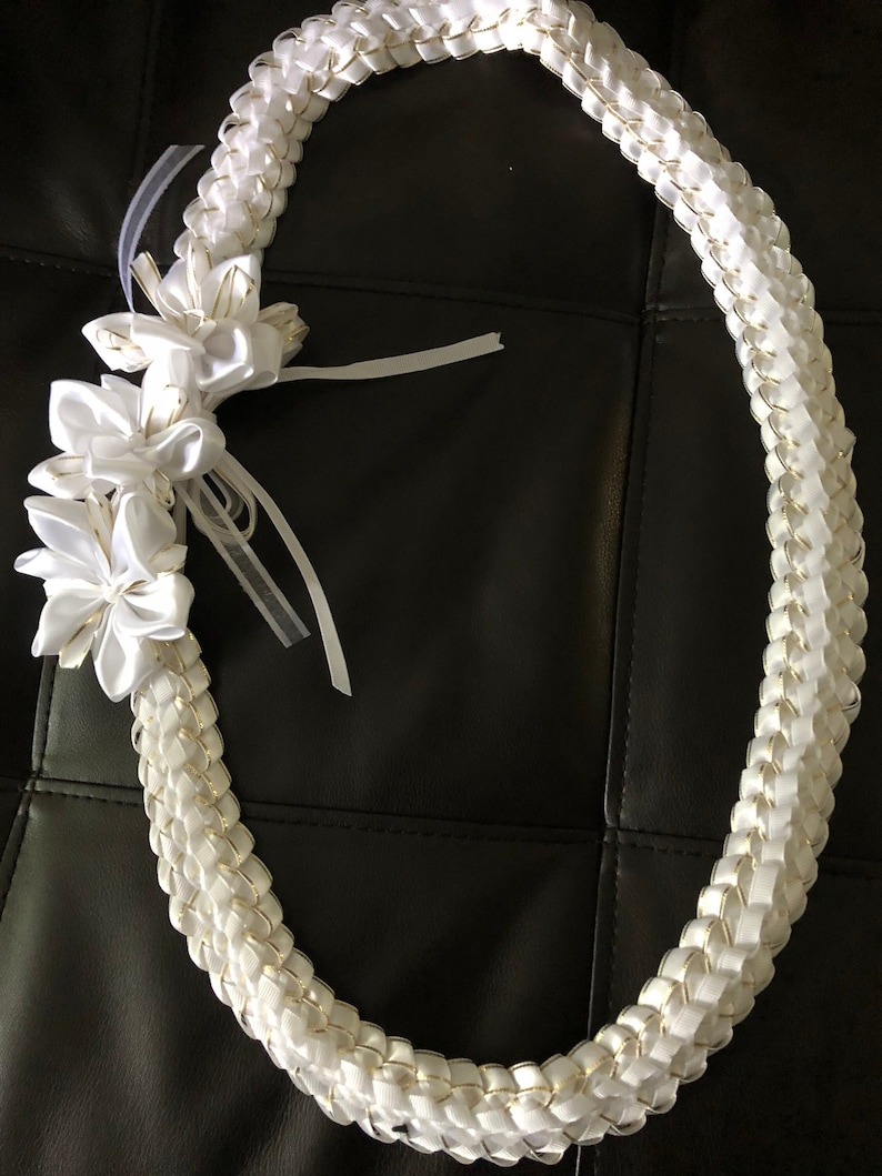 May include: A white lei necklace with a braided design, accented with gold trim. The lei features white flower embellishments and sheer ribbons. The necklace is displayed on a dark surface.