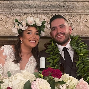 May include: A bride and groom at their wedding reception. The bride is wearing a white lace dress and a flower crown. The groom is wearing a dark suit and a lei. They are sitting at a table with flowers.