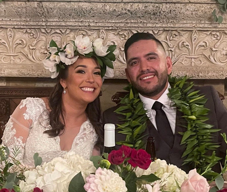 May include: A bride and groom at their wedding reception. The bride is wearing a white lace dress and a flower crown. The groom is wearing a dark suit and a lei. They are sitting at a table with flowers.
