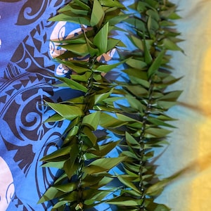 May include: A green leaf lei made of woven leaves. The lei is on a blue and black patterned fabric.