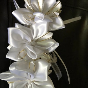 May include: A garland of white satin ribbon flowers with gold trim. The flowers are layered and have a pearl-like center. The garland is against a dark background and is suitable for weddings or other special events.