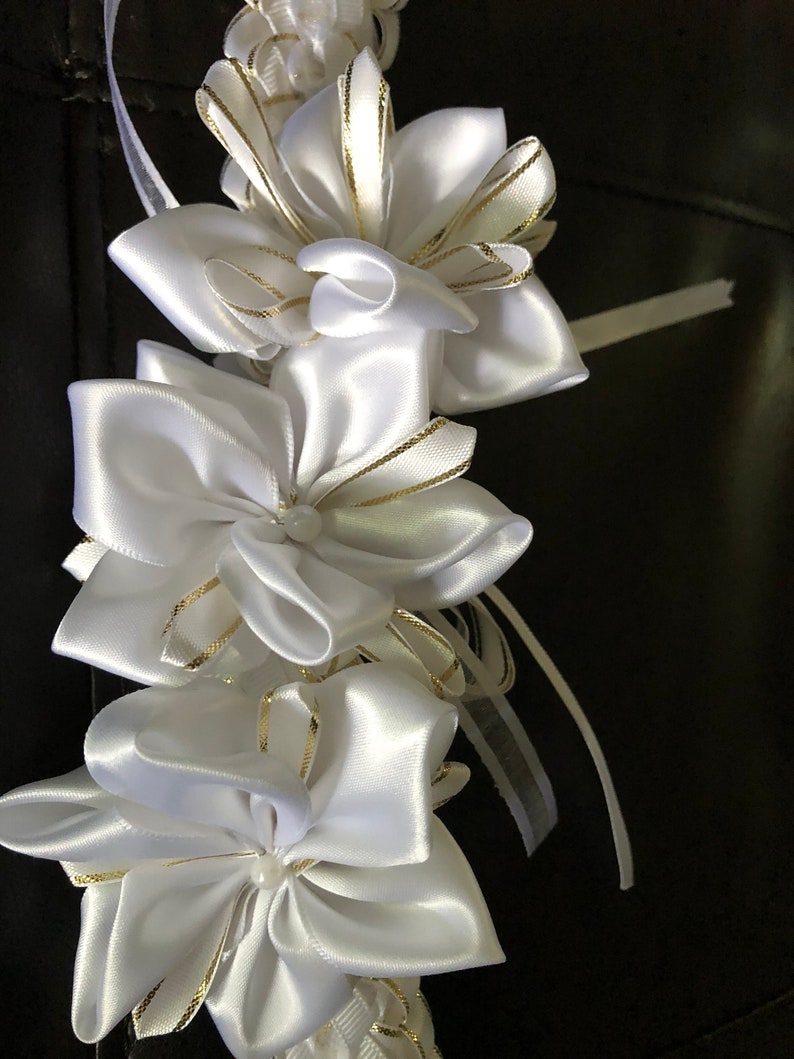 May include: A garland of white satin ribbon flowers with gold trim. The flowers are layered and have a pearl-like center. The garland is against a dark background and is suitable for weddings or other special events.
