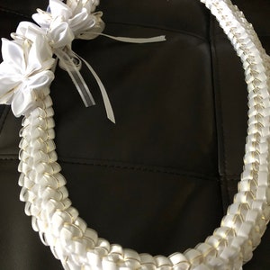 May include: A white lei with gold accents and a floral embellishment. The lei is made of woven white ribbons with gold edges, and features a large, white flower and ribbons. This decorative lei is perfect for special occasions.