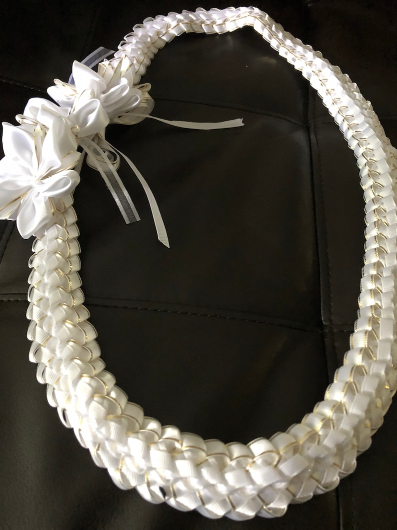 May include: A white lei with gold accents and a floral embellishment. The lei is made of woven white ribbons with gold edges, and features a large, white flower and ribbons. This decorative lei is perfect for special occasions.