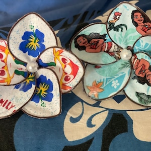 Puede incluir: Dos pinzas para el pelo de tela con forma de flor hechas a mano. Una presenta una flor azul y blanca con un personaje de dibujos animados y el texto "FAMILY". La otra presenta una flor azul y verde con un personaje de dibujos animados de la pel&iacute;cula Moana.