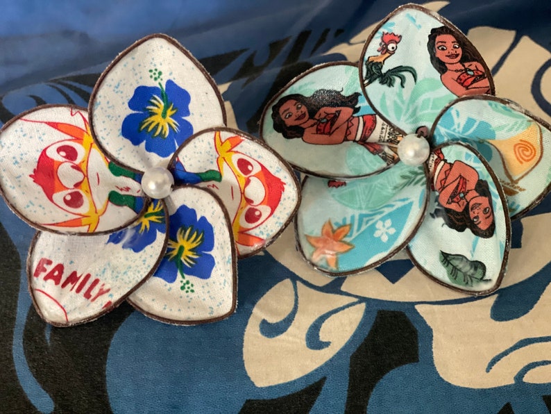 Puede incluir: Dos pinzas para el pelo de tela con forma de flor hechas a mano. Una presenta una flor azul y blanca con un personaje de dibujos animados y el texto "FAMILY". La otra presenta una flor azul y verde con un personaje de dibujos animados de la pel&iacute;cula Moana.