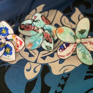 Puede incluir: Tres pinzas para el pelo de flores de tela de colores con un acento de perla blanca. Las flores presentan im&aacute;genes de la pel&iacute;cula de Disney Moana, incluidos los personajes de Moana, Maui y Pua. El texto "Ohana" y "Nadie se queda atr&aacute;s" es visible en las flores.