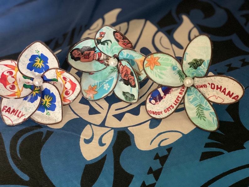 Puede incluir: Tres pinzas para el pelo de flores de tela de colores con un acento de perla blanca. Las flores presentan im&aacute;genes de la pel&iacute;cula de Disney Moana, incluidos los personajes de Moana, Maui y Pua. El texto "Ohana" y "Nadie se queda atr&aacute;s" es visible en las flores.